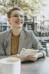 Mid adult woman looking away while reading book sitting in sidewalk cafe - MFF06714