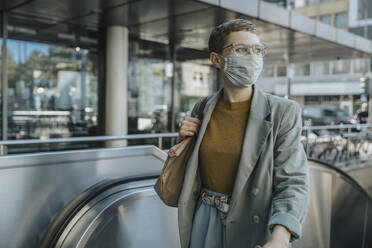 Woman wearing protective face mask looking away standing on escalator in city - MFF06715