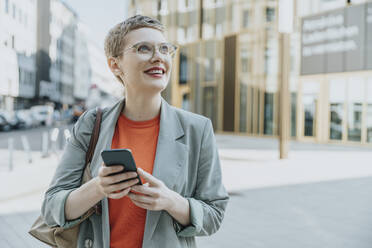 Mid adult woman looking away using smart phone standing on street in city - MFF06742