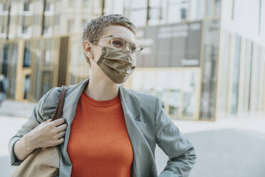 Frau mit Schutzmaske, die wegschaut, während sie an einem sonnigen Tag auf einer Straße in der Stadt steht - MFF06745