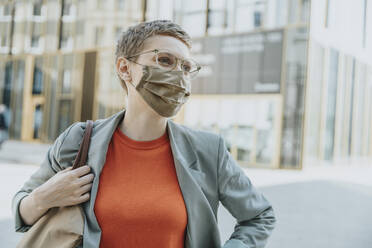 Woman wearing protective face mask looking away while standing on street in city during sunny day - MFF06745