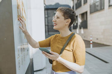 Mid adult woman reading map while holding smart phone standing in city on sunny day - MFF06760