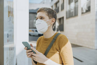 Frau mit Gesichtsschutzmaske, die an einem sonnigen Tag in einer Stadt steht und eine Karte liest, während sie ihr Smartphone in der Hand hält - MFF06761