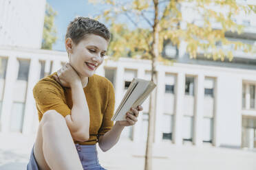 Lächelnde Frau liest ein Buch, während sie an einem sonnigen Tag auf der Straße sitzt - MFF06763