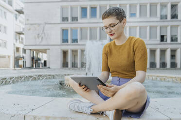 Frau im Schneidersitz mit digitalem Tablet am Brunnen sitzend an einem sonnigen Tag - MFF06769