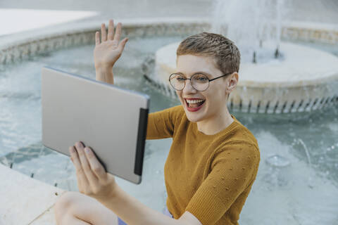 Woman doing video call with digital tablet sitting by fountain on sunny day stock photo