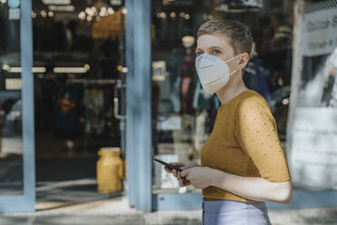 Frau mit Gesichtsschutzmaske, die ein Smartphone hält und wegschaut, während sie in der Stadt steht - MFF06775