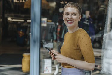 Glückliche Frau, die ein Smartphone hält und wegschaut, während sie in der Stadt steht - MFF06776