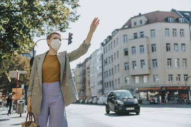 Frau mit Gesichtsschutzmaske ruft Taxi durch Winken auf der Straße in der Stadt - MFF06779