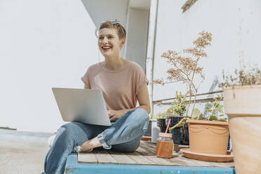 Frau lachend bei der Benutzung eines Laptops auf der Terrasse an einem sonnigen Tag - MFF06781