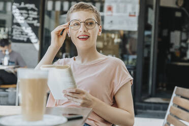 Frau, die mit einem Buch wegschaut, sitzt in einem Straßencafé an einem sonnigen Tag - MFF06790