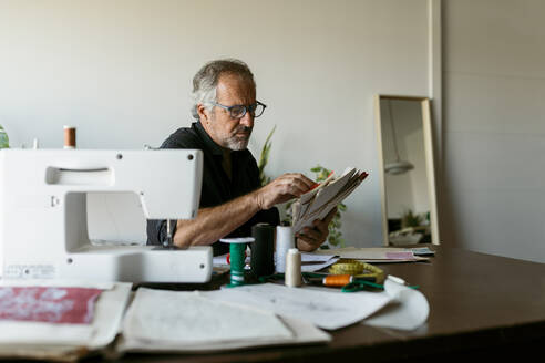 Älterer Schneider, der sich Stoffmusterpapiere ansieht, während er am Tisch im Atelier sitzt - VABF03693