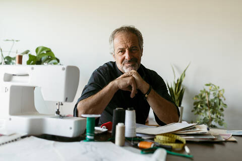 Smiling male tailor sitting at table against wall in work studio stock photo