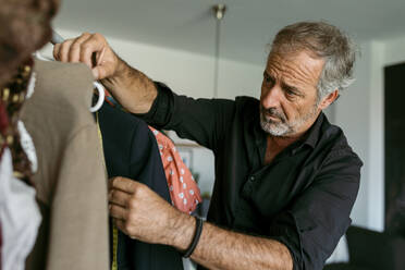 Älterer männlicher Schneider misst Kleidung auf einem Kleiderbügel im Arbeitsstudio - VABF03709