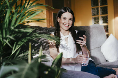 Mid adult woman using smart phone while sitting at home on sunny day stock photo