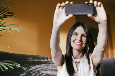 Lächelnde Frau, die ein Selfie mit ihrem Smartphone macht, während sie zu Hause sitzt - EBBF01072