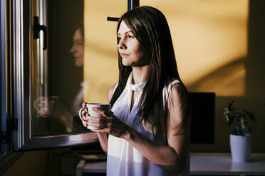 Mittlere erwachsene Frau schaut durch das Fenster, während sie mit einer Kaffeetasse zu Hause steht - EBBF01079