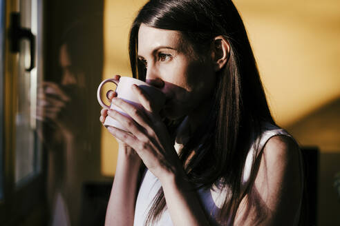 Woman looking through window while drinking coffee standing at home - EBBF01080