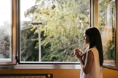 Frau schaut durch ein Fenster und hält eine Kaffeetasse, während sie zu Hause steht - EBBF01081