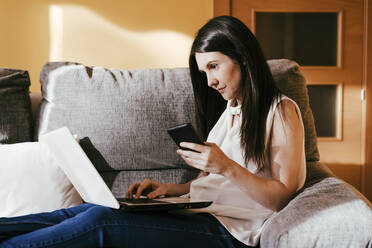 Woman using laptop and smart phone while sitting on sofa at home - EBBF01089