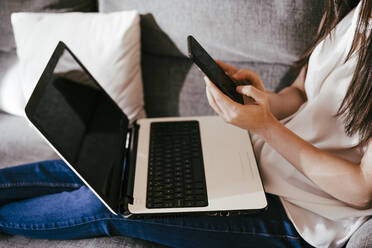 Woman using smart phone and laptop while sitting on sofa at home - EBBF01092