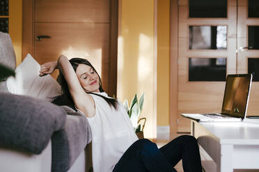 Smiling woman with hands behind head sitting with laptop at home - EBBF01095