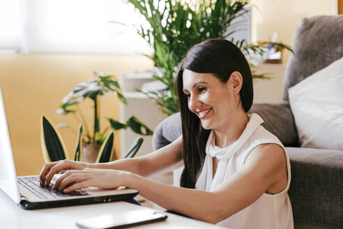 Lächelnde Frau, die zu Hause sitzend am Laptop arbeitet - EBBF01097