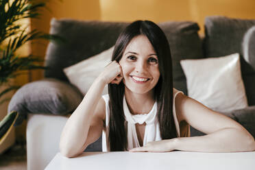 Smiling woman with hand on chin sitting at home - EBBF01098