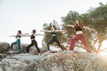 Female friends practicing yoga on rocks against clear sky during weekend - MRRF00637