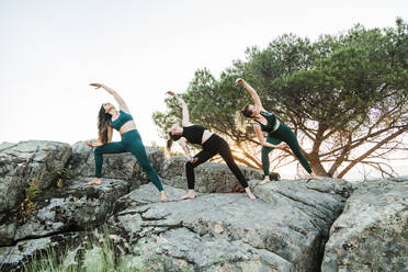 Female friends practicing yoga on rocks against clear sky during weekend - MRRF00640