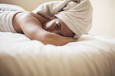 Young woman with wet hair wrapped in towel lying down on bed at home - AJOF00425