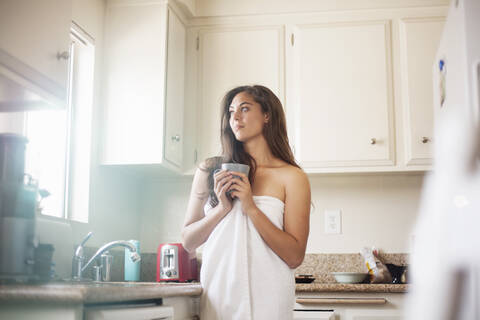 Thoughtful woman wearing towel holding coffee cup while standing in kitchen at home stock photo