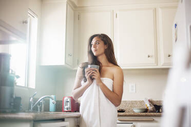 Nachdenkliche Frau, die ein Handtuch trägt und eine Kaffeetasse hält, während sie in der Küche zu Hause steht - AJOF00426