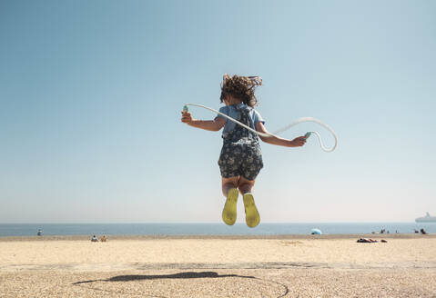 Mädchen spielt mit Springseil und springt am Strand gegen den klaren Himmel - AJOF00430