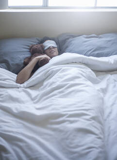 Junge Frau mit Augenmaske schläft mit Spielzeug auf dem Bett zu Hause - AJOF00432