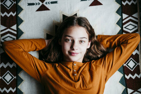 Smiling girl with hands behind head resting on blanket at home - EBBF01102