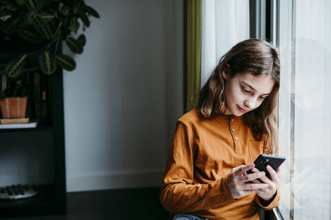 Girl using mobile phone while leaning on window at home stock photo