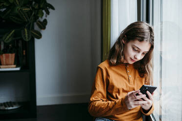 Girl using mobile phone while leaning on window at home - EBBF01105