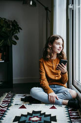 Mädchen mit Mobiltelefon, das wegschaut, während es zu Hause am Fenster sitzt - EBBF01106