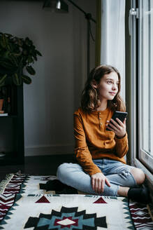 Girl with mobile phone looking away while sitting by window at home - EBBF01106