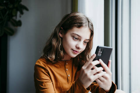 Vorpubertäres Kind, das ein Mobiltelefon benutzt, während es zu Hause am Fenster sitzt, lizenzfreies Stockfoto