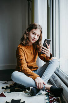 Girl with mobile phone sitting cross legged by window at home - EBBF01108