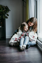 Girl sitting on sitter lap while sitting by window at home - EBBF01109