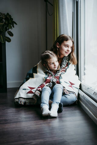 Sisters covered in blanket sitting by window at home stock photo