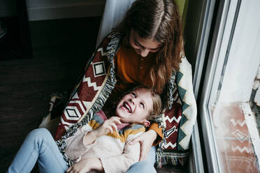 Verspieltes Mädchen, das lachend auf dem Schoß seiner Schwester zu Hause liegt - EBBF01114