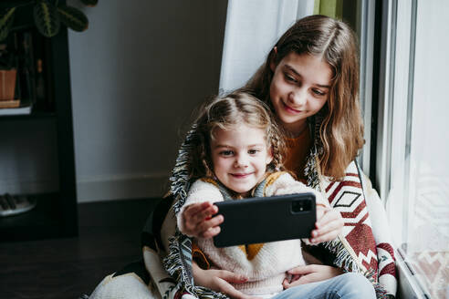 Girl taking selfie with sister while sitting by window at home - EBBF01117