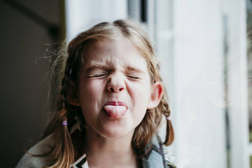 Blond girl sticking tongue out while standing by window at home - EBBF01128