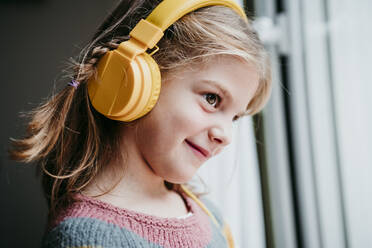 Lächelndes Mädchen mit Kopfhörern, das durch ein Fenster schaut, während es zu Hause steht - EBBF01131