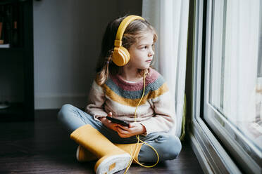 Mädchen mit Kopfhörern, das ein Mobiltelefon benutzt, während es zu Hause am Fenster sitzt - EBBF01132