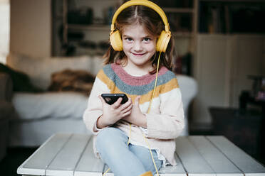 Lächelndes Mädchen mit Kopfhörern, das ein Mobiltelefon benutzt, während es zu Hause auf dem Tisch sitzt - EBBF01134